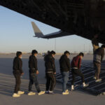 Grupo de jóvenes migrantes con mascarillas, en fila mientras abordan un avión militar en una pista de despegue.