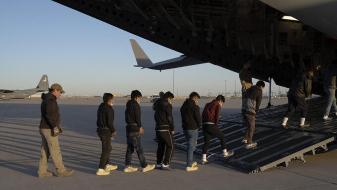Grupo de jóvenes migrantes con mascarillas, en fila mientras abordan un avión militar en una pista de despegue.