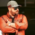 Hombre con camisa de cuero naranja y gafas oscuras, posando con los brazos cruzados frente a un coche de lujo negro.