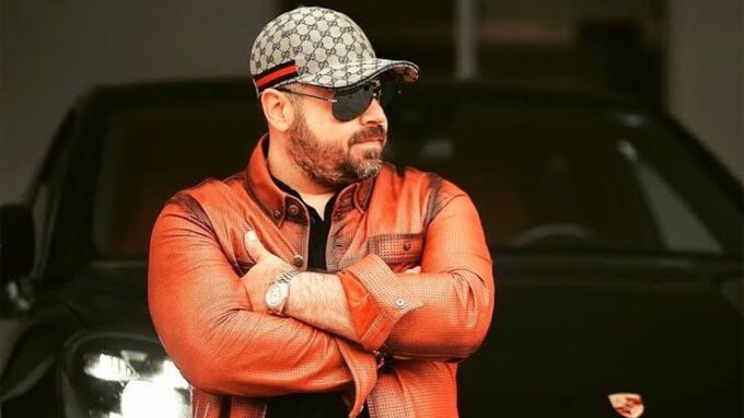 Hombre con camisa de cuero naranja y gafas oscuras, posando con los brazos cruzados frente a un coche de lujo negro.