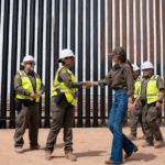 La secretaria de seguridad nacional estrecha la mano de una agente en un sitio cerca del muro fronterizo, rodeada de personal de seguridad y construcción.