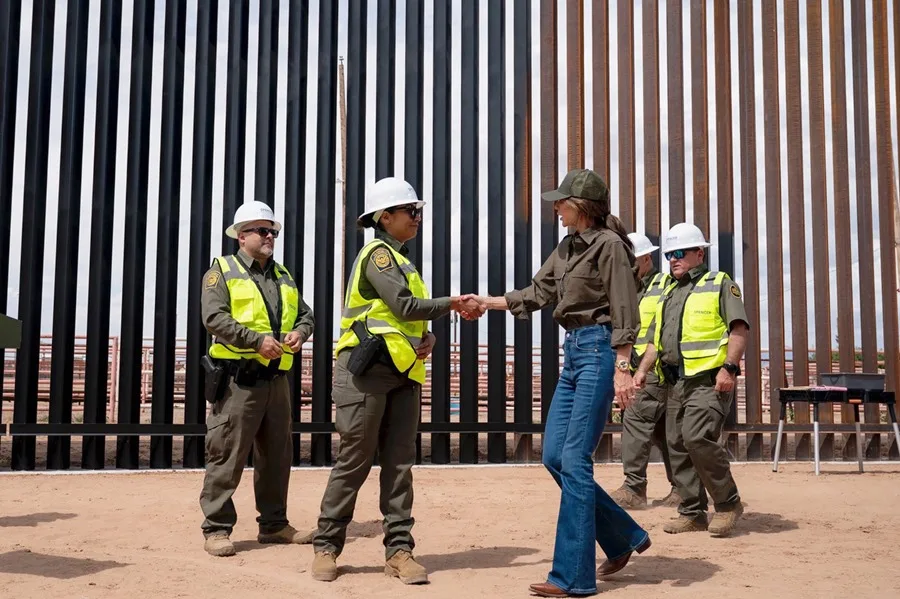 La secretaria de seguridad nacional estrecha la mano de una agente en un sitio cerca del muro fronterizo, rodeada de personal de seguridad y construcción.