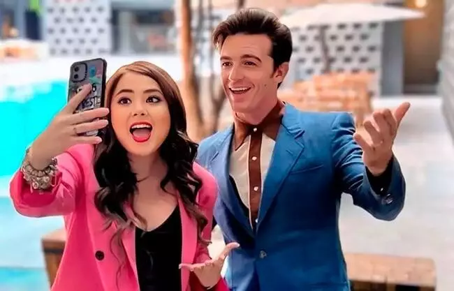 Una mujer con blazer rosa y un hombre con traje azul posan para un selfie sonrientes, con un ambiente de piscina detrás de ellos.