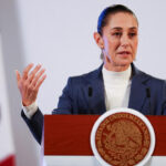 Mujer en traje azul gesticulando durante la conferencia de prensa, con la bandiera de México al fondo.